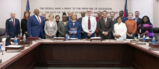 Group of diverse individuals stands around table with middle individual holding certificate
